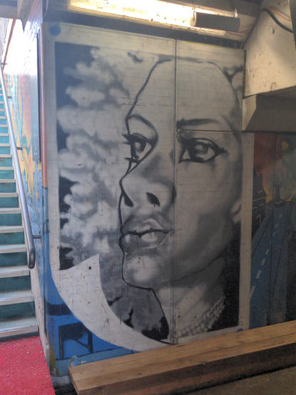 Sur un pan de mur dans un souterrain, à côté d'un escalier, un grand visage féminin de trois quarts, anguleux, en niveaux de gris.