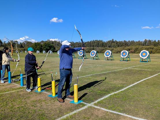 Photo of a man and boy standing together at an archery shooting line. The man is drawing his bow.