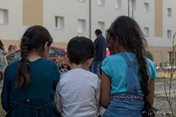 Three children are sitting with their backs to a wall. On either side are girls with plaits down their backs. In the middle, a younger boy.