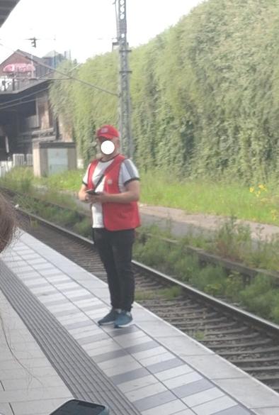 Ein Reisendenhelfer mit roter Weste und rotem Cappy steht am Bahnsteig und schaut etwas im Handy nach. Das Gesicht ist mit einem punkt unkenntlich gemacht.