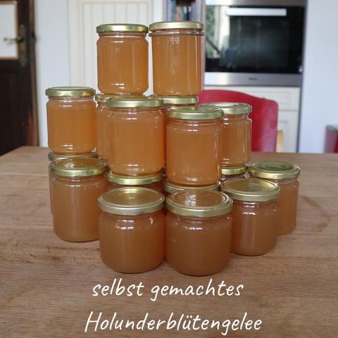 Das Bild zeigt kleine Einmachgläser, die mit frisch eingekochtem Holunderblütengelee gefüllt sind. Die Gläser sind mit goldfarbenen Metalldeckeln verschlossen und zu einer kleinen Pyramide auf einem Holztisch aufgestapelt.