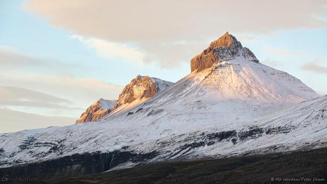 A colour photo of a mountain range at sunset, seen from the foot of the mountains. The flanks are snow- and ice-covered, with only the very tops left bare and brown. Most of the heights are in shadow, except for three sharp peaks which are highlighted with orange by the setting sun.