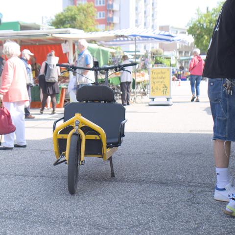 Ein Bullit Lastenrad mit Sitzbank auf der Ladefläche steht auf einem Wochenmarkt.
