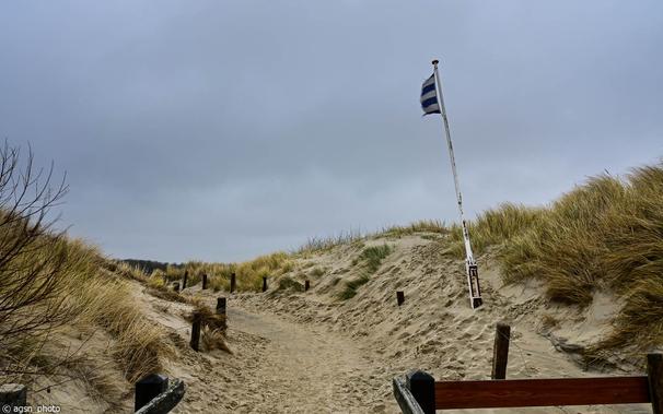 Ein Weg zwischen zwei Dünen, der Himmel ist bewölkt, eine halb zerrissene Flagge an einer Stange flattert im rauen Wind landeinwärts. / A path between two dunes, the sky is cloudy, a half-torn flag on a pole flutters inland in the rough wind.