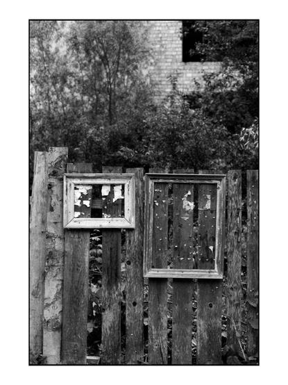 The image is a black and white photograph featuring a weathered wooden fence with two old picture frames attached to it. The frames are in a state of disrepair, with peeling paint and visible damage. The frame on the left is smaller and has a more pronounced peeling effect, while the larger frame on the right shows signs of wear with numerous small holes and remnants of paper or labels. The fence itself is made of vertical wooden planks, some of which are warped and show signs of decay. In the background, there is a brick building partially obscured by trees and foliage, adding to the sense of abandonment. The overall atmosphere of the image is one of neglect and the passage of time.