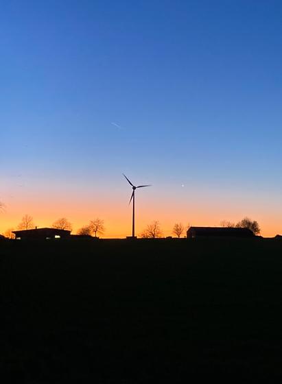 Eine Sonnenuntergangsszenerie. Oben noch blauer Himmel, dann ein Strefen orange und im Vordergrund ein fast schwarzes Feld. Am Horizont zentral in der Mitte ein Windrand, rechts und links einzelne kahle Bäume und zwei Hofgebäude.