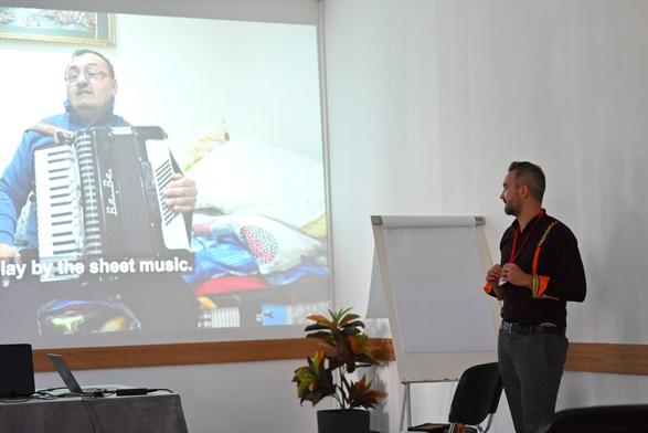 A man presenting a video with an accordionist at a conference setting.