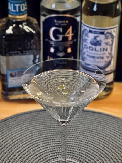 clear cocktail with olive oil drops as garnish in a chilled coupe with bottles of G4 108 proof blanco tequila, Olmeca Altos plato tequila, and Dolin vermouth blanco in the background