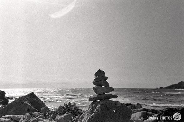 A black and white photograph of a pile of stacked pebbles on a rock against the sea and a clear sky