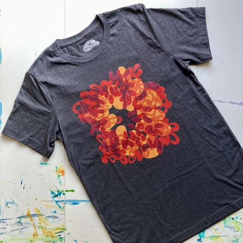 Dark heather grey short-sleeved t-shirt laid out flat on a white tabletop that has dried smears and drips of paint on it. On the front of the t-shirt is printed an abstracted fire burst with an empty centre, like a wriggly and swirling doughnut of red, orange, and gold yellow overlapping shapes.