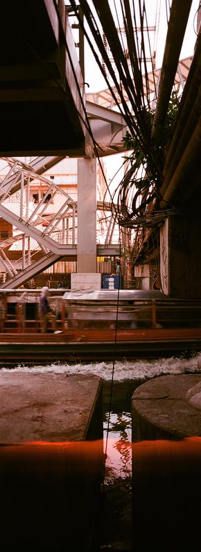 EN: vertical panoramic photo in color with very strong red tints of a blurred boat passing in front of concrete installation and under a tangle of electrical wire. On the other side, a complex staircase structure rises above the canal

FR: photo panoramique format portrait, en couleur avec des rouges très marqués, dd'un bateau flou qui passe devant des structures en béton et sous un enchevêtrement de fils électriques. De l'autre côté s'élève une straucture assez complexe qui est en fait un escalier