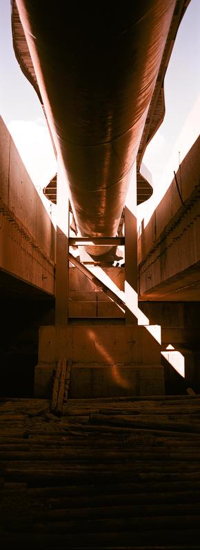 EN: vertical panoramic photo in color with very strong red tints of a pipe between two bridges, take from below. Below the pipe and bridges is concrete and below that, wood resting in the darkness

FR: photo panoramique format portrait, en couleur avec des rouges très marqués, d'un tuyau entre deux ponts, pris du dessous. En bas, du béton, et encore en dessous, des rondins de bois dans la pénombre