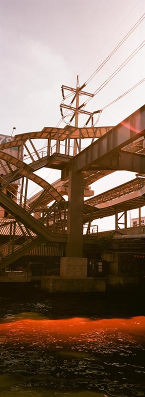 EN: vertical panoramic photo in color with very strong red tints of a complex metal and concrete structure on the other side of the canal, including an electric post, metro rails and a staircase. Light has leaked on the bottom, giving the water a weird red extra wave

FR: photo panoramique format portrait, en couleur avec des rouges très marqués, d'une structure complexe en métal et en béton de l'autre côté du canal, comprenant des rails de métro aérien, un poteau électrique et un escalier. En bas, la lumière a fui, ce qui fait une nouvelle vague rouge bizarre sur l'eau