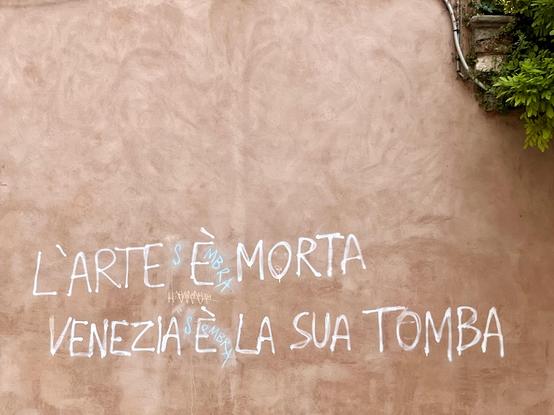 A pastel-red wall, with a bit of greenery visible in the top right hand corner of the frame. Someone has graffitied in white capital letters, "l'arte è morta, Venezia è la sua tomba" (Art is dead, Venice is its tomb) to which someone has added in pale blue an "s" and "mbra" on either side of the e, so it now reads, much more accurately, "L'arte sembra morta, Venezia sembra la sua tomba" (Art appears dead, Venice appears to be its tomb)