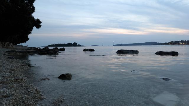 Blick aufs Meer, im Wasser sind ein paar Steine die über die Oberfläche ragen, links Kiesstrand und ein Baum, im Hintergrund das gegenüberliegende Ufer und eine Insel
