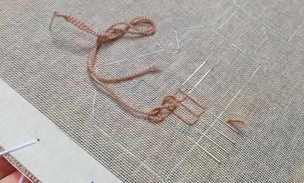 The beginning of a stitched artwork on an open weave fabric. Tacked outlines resemble a house and a window frame has been stitched in brown wool. Pins have been used to indicate where to stitch