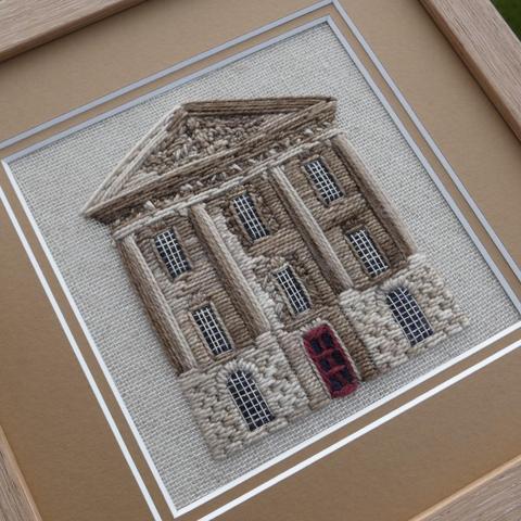 A framed stitched artwork depicting the front, middle section of Chatsworth House. Four stone pillars hold up a sculpted portico. Eight multipaned windows on three floors with a red door central on the ground floor.