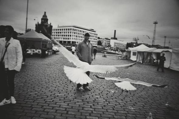 This is a landscape-orientated black and white, eye-level, medium shot of a cobblestone square where two large birds are captured mid-flight, seemingly taking off or landing. In the foreground, a person stands on the left, partially cropped out, wearing a light-colored jacket and dark pants, with white sneakers. In the middle ground, another person, wearing a dark cap and a light-colored jacket over dark pants, stands facing the camera. Behind them, several large tents are set up, and in the background, multi-story buildings and a church steeple are visible under an overcast sky. Other indistinct figures are present in the distance.