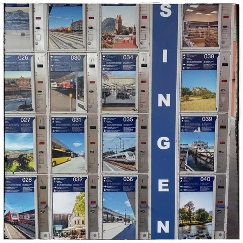 Die Gepäckaufbewahrungsboxen in Singen sind mit Bildern beklebt. Meist Bahnhöfe, aber auch natur. Das quadratische Foto mit wrißem Rahmen und den im Raster gesetzten Bildern sieht aus wie eine zu lösende Captcha-Frage. Senkrecht geschrieben steht Singen im rechten Drittel des Bilds.