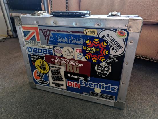 An old tour briefcase made by Anvil Cases of Rosemead, California. It's covered in stickers advertising everything from synthesizers to theme parks, from ski resorts to surf wear, and record labels to effects pedals. You can just about make out that it was originally blue. It's sitting on the floor of Chris's living room.