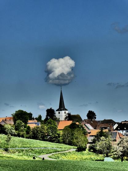 Wir schauen auf die Kirche von Ottenstein, dem Hauptort der Ottensteiner Hochebene im Weserbergland.  Der Himmel ist fast blau und eine große Wolke scheint auf der Spitze des Kirchturms zu balancieren, ihre Unterseite ist graublau, die Oberseite weiß.