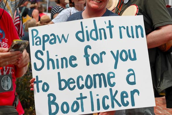 Image is of the "No Kings" protest in Abingdon, VA on 6/14/25.

A person holds a white sign with blue lettering. The sign reads: "Papaw didn't run Shine for you to become a Bootlicker".