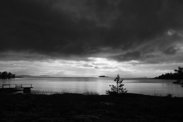Black-and-white photograph of a Finnish archipelago seascape at the end of the day at Midsummer. There are dark storm clouds dominating the sky with the clouds clearing and the sun breaking through near the horizon. There are silhouettes of islands in the distance in the centre of the frame and on the left and right sides of the photo. The island on which the photographer is standing is also silhouetted - with shadows of a pier and a pine tree silhouetted against the ripply sea. West coast of Finland, 19 June 2015. Photo Cindy Kohtala
