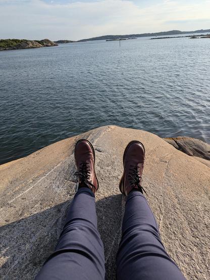 Mes jambes étendues devant moi alors que je suis assise sur un gros rocher beige et plat en bord de mer. Je porte un pantalon bleu-violet et des Doc Martens rouge cerise. La mer est plate, le ciel bleu légèrement voilé, on aperçoit des îles boisées à l'horizon.