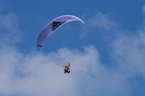 A paraglider against a deep blue sky with some light clouds in the lower half of the background. The wing is mostly white, the pilot's harness, outfit and helmet are a mixture of black and orange. The pilot is looking away from the camera, into the direction he's presumable turning to.