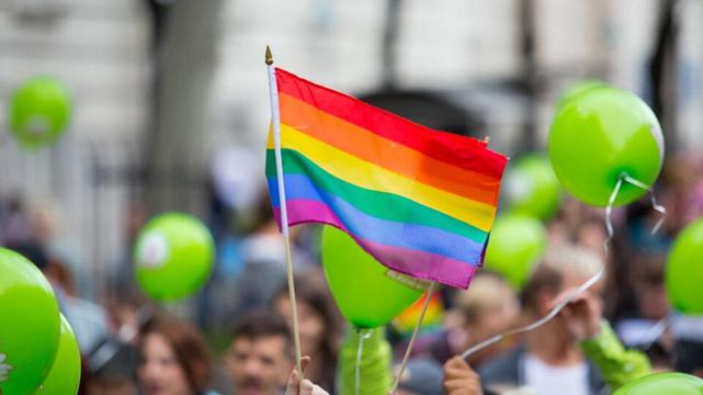 Regenbogenfahne weht bei einer Pride-Demonstration zwischen grünen Luftballons. Im Hintergrund eine Menschenmenge, unscharf, in Bewegung.