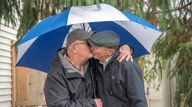 Zwei ältere Männer küssen sich liebevoll unter einem blau-weiß gestreiften Regenschirm. Beide tragen Jacken und Schirmmützen. Im Hintergrund sind grüne Tannenzweige und ein Holzzaun zu sehen.