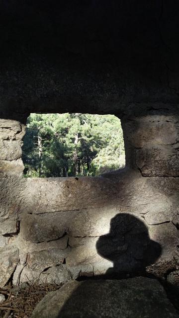 A lookout or sniper post looks out onto a hillside. It is a stone wall with a square hole. A shadow showing the outline of my head and shoulders appears on the wall.