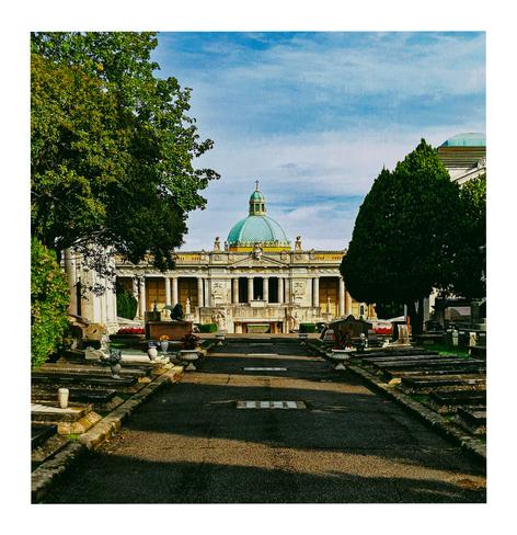 The photo is of a long, straight path in a cemetery that leads towards a large, ornate building with a teal coloured dome. The building appears to be a mausoleum or ossuary, with classical architectural features like columns and statues. On either side of the path are rows of gravestones on the ground in the grass. On both sides of the photo green trees frame the scene.