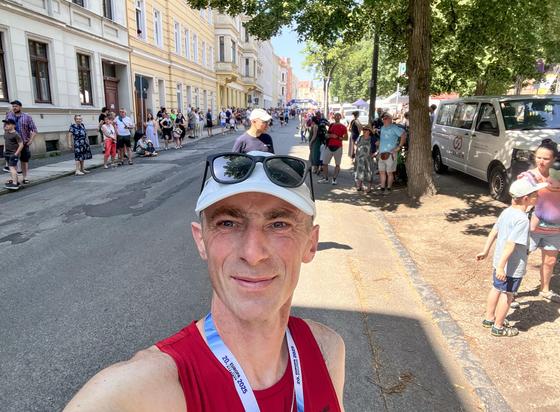 Mężczyzna w czerwonym podkoszulku i okularach przeciwsłonecznych robi selfie na słonecznej ulicy. Tłum ludzi jest widoczny w tle, wraz z drzewami wzdłuż drogi i zaparkowanymi pojazdami. Scena wydaje się być żywa i świąteczna.