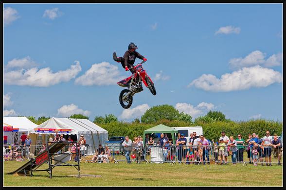 A motorcyclist performs a mid-air stunt after launching off a ramp labeled "STUNT RIDER." Wearing a helmet and protective gear, they maneuver a red motorcycle adorned with decals. A crowd watches from behind a metal barrier, with tents and a green canopy visible in the background. The bright blue sky with scattered clouds adds to the dramatic action of the scene.