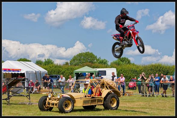 A motorcyclist launches off a ramp, soaring over a yellow off-road vehicle with the license plate "HAY 150." The red dirt bike is mid-air against a backdrop of blue sky and scattered clouds. Spectators watch from behind a metal barrier, with some capturing the stunt on cameras. The setting is an outdoor event on a grassy field, with tents, vehicles, and a white van visible in the background.
