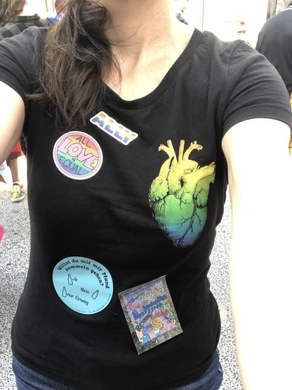 Das Bild zeigt die Oberkörper eines Menschen, der ein schwarzes T-Shirt trägt, das mit verschiedenen Aufklebern verziert ist. Auf der linken Brustseite befindet sich ein Aufkleber mit dem Text "ALL LOVE IS EQUAL" in bunten Farben. Darüber ist ein kleinerer Aufkleber mit dem Wort "ALLY" in den Regenbogenfarben zu sehen. Rechts auf dem T-Shirt ist ein großes Herz in einem Farbverlauf von Gelb, Grün und Blau abgebildet. Unten links auf dem T-Shirt befindet sich ein blauer Aufkleber mit der Frage "Willst du mit mir pfand sammeln gehen?" und den Antworten "Ja" und "Nein nur Einweg". Der Hintergrund ist unscharf und zeigt andere Personen und ein gepflastertes Bodenstück.