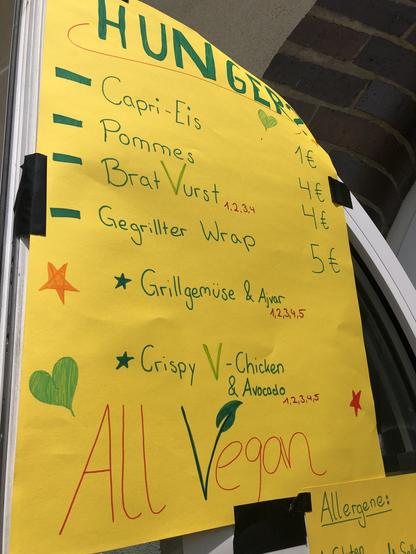 Das Bild zeigt ein gelbes Papier mit einer Handgeschriebenen Menüliste, die "HUNGERZ" lautet. Die Menüpunkte sind in grüner und roter Schrift aufgelistet und umrandet mit Zeichen wie Herzen und Sternen. Die Preise sind in Euro angegeben. Die Menüpunkte sind:

Capri-Eis: 1€
Pommes: 4€
Brat Vurst: 4€
Gegrillter Wrap: 5€
Grillgemüse & Ajvar
Crispy V-Chicken & Avocado
Unter dem Menü steht "All Vegan" in roter Schrift. Am unteren Rand des Papiers ist ein kleiner Text mit "Allergene:" zu sehen, aber die genauen Allergene sind nicht sichtbar. Das Papier ist mit Klebeband an einer Wand befestigt, die aus Ziegeln besteht.
