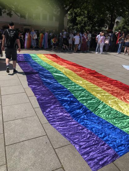 Das Bild zeigt eine große, bunte Regenbogenflagge, die auf einem gepflasterten Platz ausgebreitet ist. Die Flagge besteht aus sechs horizontalen Streifen in den Farben Rot, Orange, Gelb, Grün, Blau und Violett. Im Hintergrund ist eine Gruppe von Menschen zu sehen, die sich um die Flagge versammelt hat. Einige Personen tragen Regenbogenkleidung, was auf eine LGBTQ+-Bewegung hinweist. Die Atmosphäre wirkt friedlich und festlich. Ein Mann in einem schwarzen T-Shirt und schwarzen Shorts geht an der Flagge vorbei. Die Sonne scheint, und es sind Bäume im Hintergrund zu sehen, die Schatten auf den Platz werfen.
―