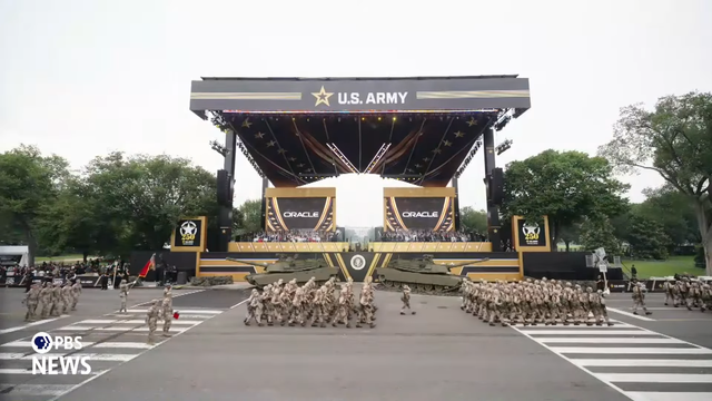Trumps Military Parade on PBS News. Military personnel marching in front of two tanks on display and a giant U.S. Army stage. On the stage are two giant screens showing the Oracle logo.