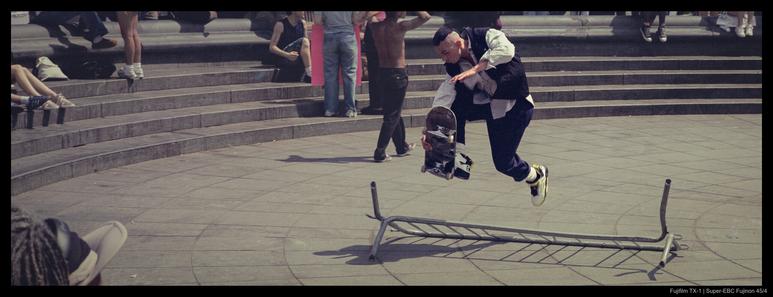 A skateboarder performs a trick jump over a toppled metal barrier in a dry fountain bed.