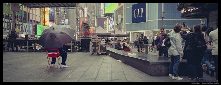 A person beneath an umbrella sits in a red metal chair, separated by a large stone bench and empty space from a crowd of pedestrians in Times Square.