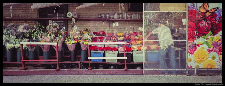A person stands by a large shelf of flowers in an outdoor flower shop, mounted to the side of a building.  A bright decorative image of flowers is painted on one partition.