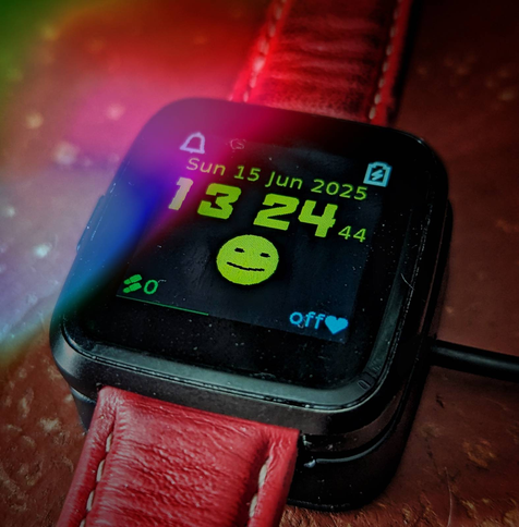 A pine time smart watch showing the time at nearly half past one on a Sunday, and a smiley face which is now smiling slightly. The charging icon indicates it is charging up.