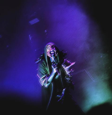A performer with a textured, possibly masked, face and dark, hooded attire stands on a stage bathed in blue and green spotlights, holding a microphone.