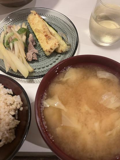 A plate containing fried zucchini, sautéed vegetables, and thinly sliced meat, next to a bowl of miso soup with cabbage. Brown rice is served on the side, along with a glass of chilled beverage.