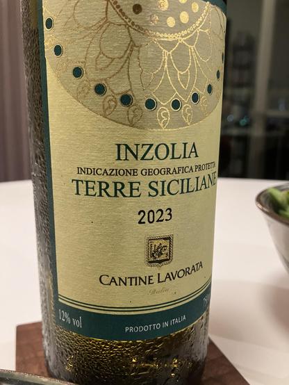 Close-up of a wine bottle labeled "Inzolia Terre Siciliane 2023" from Cantine Lavorata, featuring decorative design elements. The bottle has a 12% alcohol content and is produced in Italy. A bowl of salad