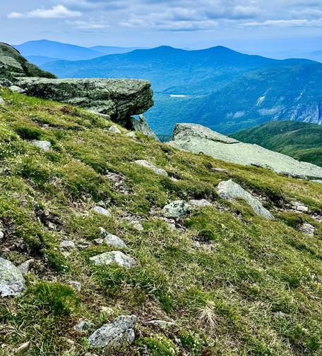 Photo of distant mountains as viewed from steep slope of another mountain. Grass in foreground. Steep slope shown angling down from left to right. A large angular boulder at intermediate distance on slope. Distant mountains in background behind the boulder with several different peaks at different distances. Cloudy skies overhead.

I took this photo on my backpacking trip several days ago as I was descending from Mt Lafayette. The view is toward the south.