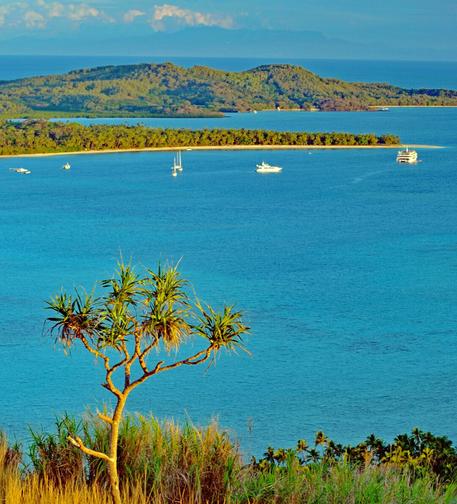 Photo shows an outlook view from an elevated hill towards a set of smaller pacific islands. The nearest is a low crescent shaped one with palm trees and forest and a white beach. Behind the low island is an island with a set of small hills and small natural harbours with some huts and some boats moored. Behind the Pacific is stretching towards the horizon where there might be some island shapes under a light blue afternoon sky, with some clouds building up. In front of the photo there is a single small tree sharing the view. Down the hill for the tree, there is a larger body of water and two thirds of the way to the low beach island there are a collection of sailboats, motorboats and a small cruise ship berthed for the night. The sea is deep indigo blue. The tree has the warm colours of a late afternoon setting sun.