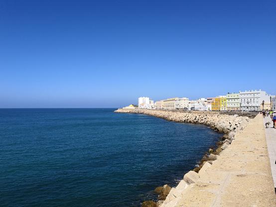 Vista del malecón de Cádiz con gente paseando por él. A la izquierda se puede ver el Océano Atlántico.

View of the Cadiz boardwalk with people walking through it. On the left you can see the Atlantic Ocean.
